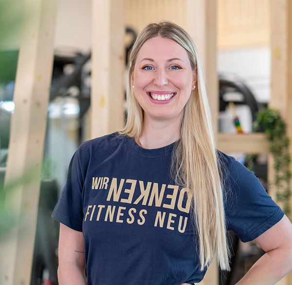 Jeannine mit langem blondem Haar lächelt, trägt T-Shirt mit Aufdruck "Wir denken Fitness neu‘“