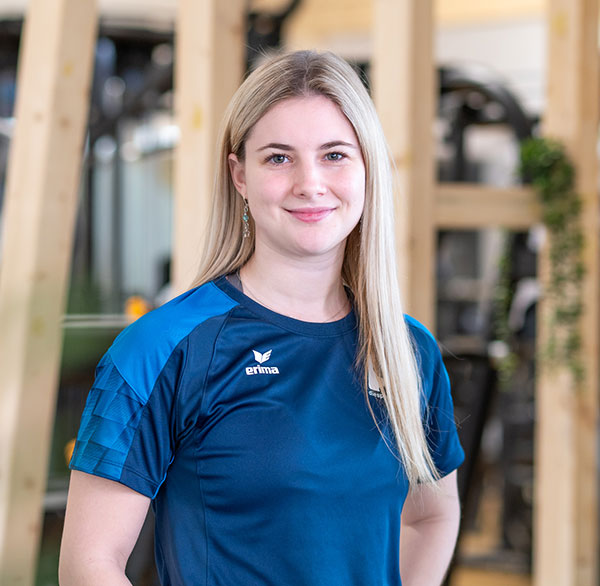 Junge Frau in Sportshirt lächelt leicht, steht in modernem Trainingsbereich mit Holzelementen