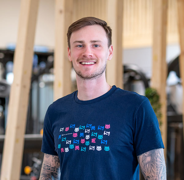 Junger Mann mit Tattoos und T-Shirt mit Superheldenmotiv lächelt in hellem Trainingsraum