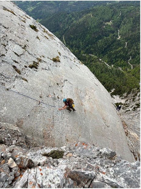 Kletterin mit Helm an steiler Felswand, Blick ins grüne Tal im Hintergrund
