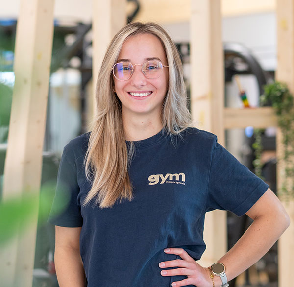 Junge Frau mit Brille und blondem Haar lächelt, trägt dunkles T-Shirt mit ‚gym  diesportpraxis‘