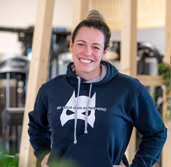 Frau im Hoodie mit Superhelden-Logo lächelt, steht im Trainingsraum mit Holzelementen