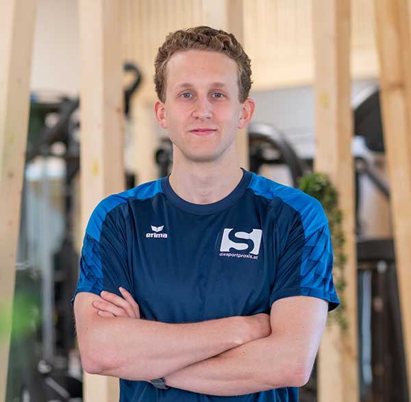 Junger Mann in Sportkleidung mit verschränkten Armen steht in einem Fitnessstudio und blickt selbstbewusst in die Kamera.